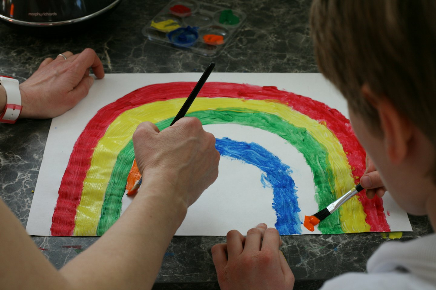 Mani di adulto e bambino dipingono un arcobaleno su carta bianca.